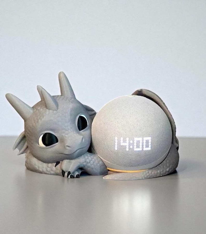 May include: A gray dragon-shaped speaker holder cradling a spherical speaker. The speaker displays the time, 14:00, in white digits. The dragon has large black eyes and a friendly expression.