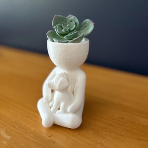 Puede incluir: Una maceta de cer&aacute;mica blanca con forma de persona que sostiene un perro, con una planta suculenta en la cabeza. La figura est&aacute; sentada con las piernas cruzadas. La suculenta tiene hojas verdes. Fondo: pared azul oscuro y mesa de madera.
