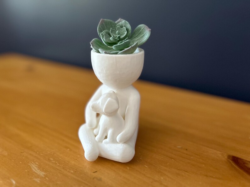 Puede incluir: Una maceta de cer&aacute;mica blanca con forma de persona que sostiene un perro, con una planta suculenta en la cabeza. La figura est&aacute; sentada con las piernas cruzadas. La suculenta tiene hojas verdes. Fondo: pared azul oscuro y mesa de madera.
