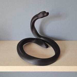 以下が含まれることがあります： 黒いヘビの彫刻。ヘビの頭は持ち上がり、体はループ状になっています。彫刻は、灰色の壁を背景に、明るい色の木製の棚に置かれています。ヘビはマットブラックです。