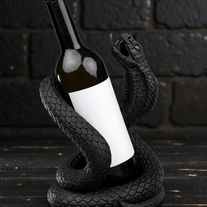 Puede incluir: Un soporte negro para botellas de vino con forma de serpiente enroscada alrededor de una botella de vino. La serpiente tiene escamas detalladas y la boca abierta. La botella tiene una etiqueta blanca en blanco.