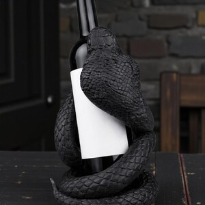 Puede incluir: Una botella de vino negra sujeta por un soporte decorativo de vino con forma de serpiente negra. El cuerpo de la serpiente se enrolla alrededor de la botella, creando una presentaci&oacute;n &uacute;nica y elegante. La botella tiene una etiqueta blanca.