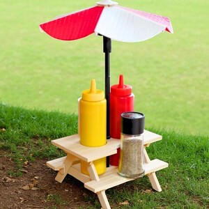 Pode incluir: Uma pequena mesa de piquenique de madeira com um guarda-sol às riscas vermelhas e brancas. Na mesa estão garrafas de mostarda amarela e ketchup vermelho, além de um pimenteiro de vidro. A mesa está na relva.