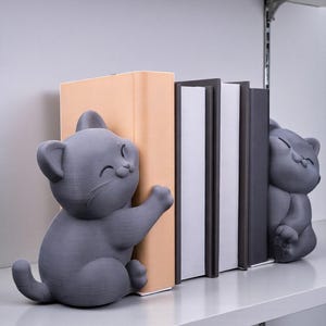 May include: Gray cat-shaped bookends. One cat is hugging a book, the other is sitting. The bookends are on a white shelf with books between them. The books have a light tan, white, and dark gray color scheme.