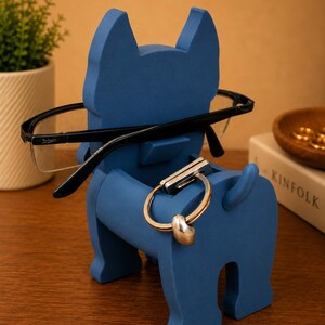 May include: A blue dog-shaped eyeglasses holder with black-framed glasses resting on the nose. A silver and gold key ring hangs from the dog's body. A small plant and a book are in the background.