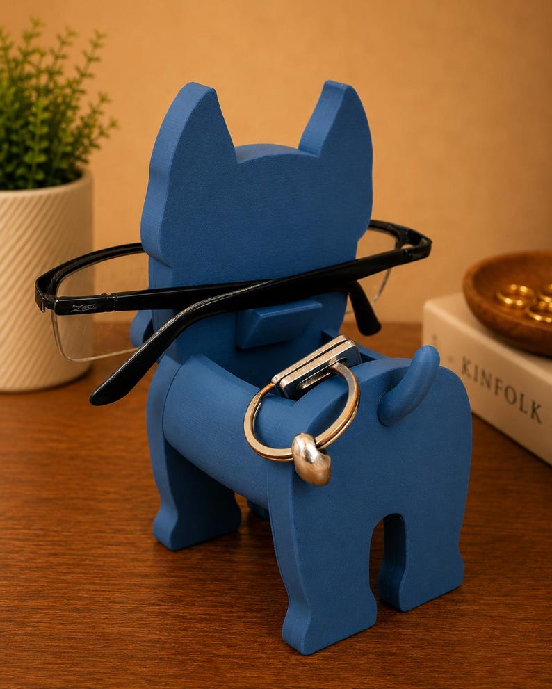 May include: A blue dog-shaped eyeglasses holder with black-framed glasses resting on the nose. A silver and gold key ring hangs from the dog's body. A small plant and a book are in the background.