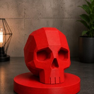 May include: A red, geometric skull sculpture. The skull is displayed on a matching red circular base. The sculpture has a faceted design, with visible planes and angles. The background includes a plant and a decorative light fixture.