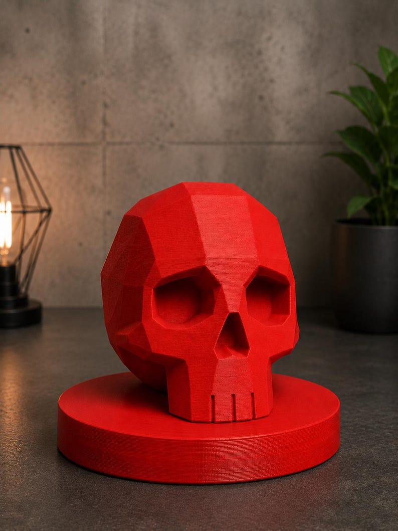 May include: A red, geometric skull sculpture. The skull is displayed on a matching red circular base. The sculpture has a faceted design, with visible planes and angles. The background includes a plant and a decorative light fixture.
