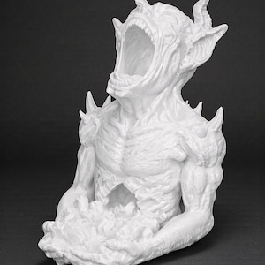 May include: A white sculpture of a creature with an open mouth, horns, and sharp protrusions. The figure has muscular arms and is holding a mass of undefined material. The sculpture is set against a dark background.