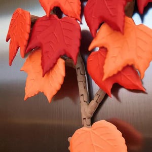 May include: A decorative fall-themed piece featuring a branch with vibrant, textured leaves. The leaves are in shades of red, orange, and yellow, creating a warm, autumnal aesthetic. The branch is a light brown color.