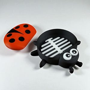 May include: A whimsical ladybug-themed soap dish and lid set. The dish is black with a white, slatted interior, and large cartoon eyes. The lid is orange with black spots, mimicking a ladybug's shell. A fun bathroom accessory.