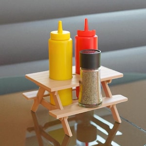 May include: A miniature picnic table holds yellow mustard, red ketchup, and a glass pepper grinder. The condiments are in squeeze bottles. The table is made of light-colored wood.