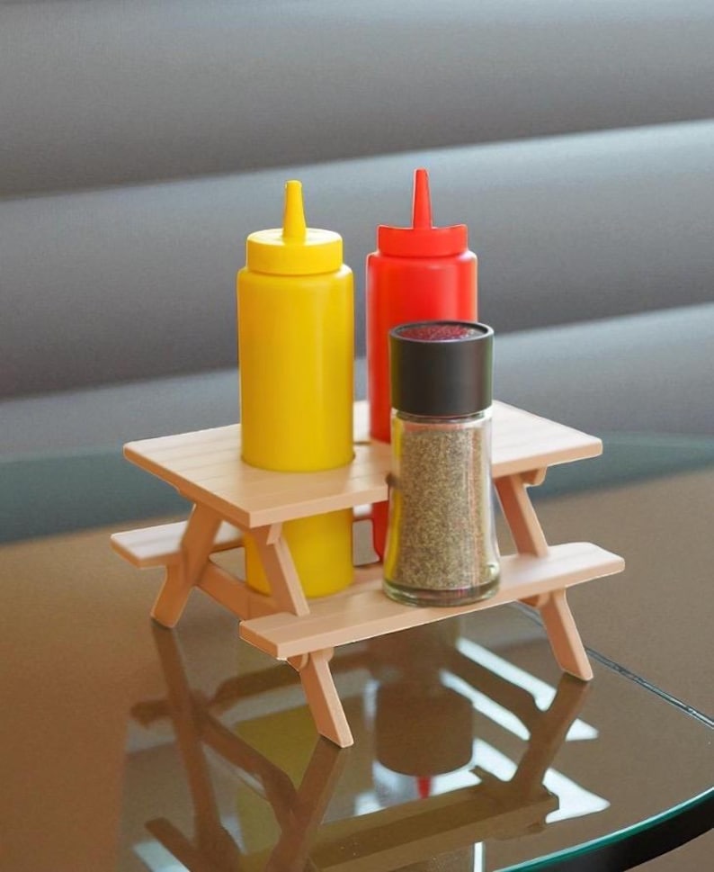 May include: A miniature picnic table holds yellow mustard, red ketchup, and a glass pepper grinder. The condiments are in squeeze bottles. The table is made of light-colored wood.