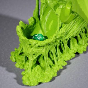 May include: A bright green, organic-looking dice holder with a single green and white die inside. The holder has a textured, uneven surface with a small opening to hold the die. The overall design is reminiscent of a plant or organic form.