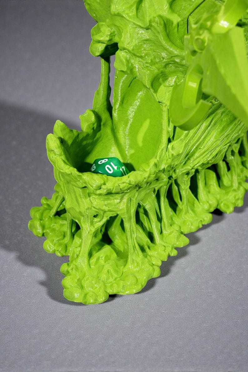 May include: A bright green, organic-looking dice holder with a single green and white die inside. The holder has a textured, uneven surface with a small opening to hold the die. The overall design is reminiscent of a plant or organic form.