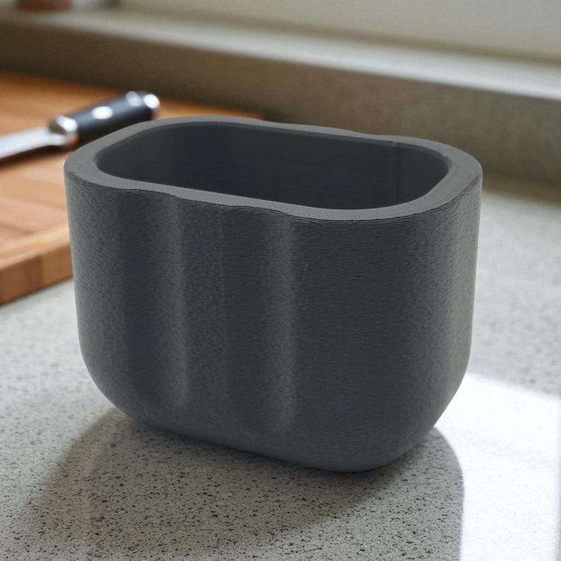May include: A dark gray, rectangular container with a textured surface. The container has a rounded bottom and a wavy design on the sides. A knife and cutting board are visible in the background.