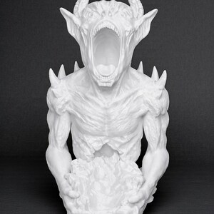 May include: A white sculpture of a demonic creature with an open mouth, horns, and sharp protrusions. The figure is holding a textured mass in its hands, set against a dark background. The sculpture has a detailed, textured surface.