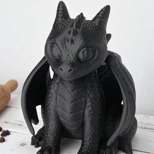 May include: A black dragon figurine with large wings, sitting upright. The dragon has large, round eyes and detailed scales. The figurine is made of a matte black material and is set against a light-colored background.