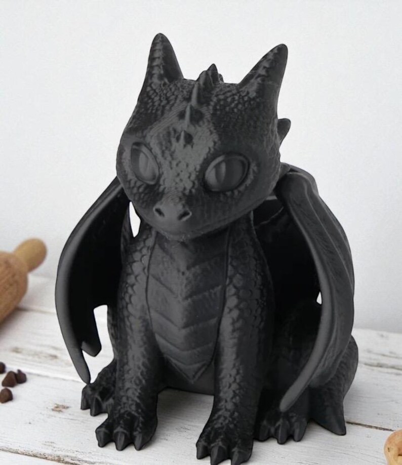 May include: A black dragon figurine with large wings, sitting upright. The dragon has large, round eyes and detailed scales. The figurine is made of a matte black material and is set against a light-colored background.