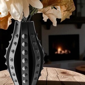 Puede incluir: Un jarr&oacute;n decorativo negro y gris con un dise&ntilde;o &uacute;nico de tachuelas, que contiene flores blancas y naranjas y follaje de oto&ntilde;o. El jarr&oacute;n est&aacute; hecho de un material oscuro y se encuentra sobre una superficie de madera. En el fondo se ve una chimenea.
