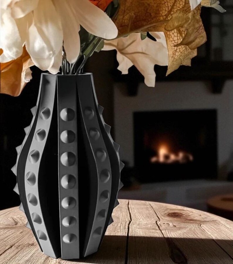Puede incluir: Un jarr&oacute;n decorativo negro y gris con un dise&ntilde;o &uacute;nico de tachuelas, que contiene flores blancas y naranjas y follaje de oto&ntilde;o. El jarr&oacute;n est&aacute; hecho de un material oscuro y se encuentra sobre una superficie de madera. En el fondo se ve una chimenea.