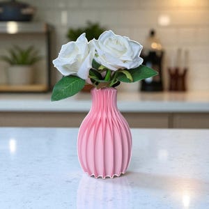 May include: A pink vase with a pleated design holds two white roses with green leaves. The vase is made of a solid material and has a rounded shape. The flowers are in full bloom, adding a touch of elegance to the arrangement.
