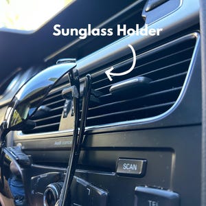 May include: A black sunglass holder attached to the air vent in a car. The holder is made of two clips that hold the sunglasses in place. The text "Sunglass Holder" is written above the holder.