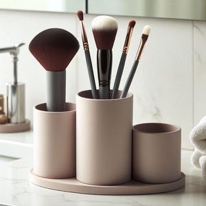 Op de afbeelding: Een blushkleurige make-up borstelhouder met drie cilindrische compartimenten en een bijpassende ovale basis. De houder bevat verschillende make-up borstels met zwarte, grijze en gouden handvatten en bruine en witte borstelharen. De houder staat op een wit aanrecht.