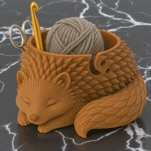 May include: A brown hedgehog-shaped yarn bowl with a ball of gray yarn inside. A gold crochet hook and silver scissors are placed in the bowl. The hedgehog has a textured, spiky design and is resting on a dark surface.
