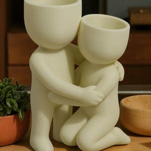 K&ouml;nnte beinhalten: Ein cremefarbener Keramik-Blumentopf in Form von zwei sich umarmenden Figuren. Eine Figur steht, w&auml;hrend die andere kniet, die Arme umeinander gelegt. Die K&ouml;pfe der Figuren sind offen und bilden Pflanzt&ouml;pfe. Im Hintergrund ist eine kleine Pflanze zu sehen.