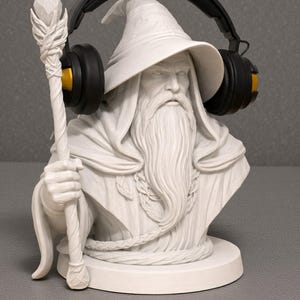 May include: A white bust of a wizard with a long beard, holding a staff, and wearing black headphones with gold accents. The wizard is wearing a hat and a robe. The bust is on a round base.