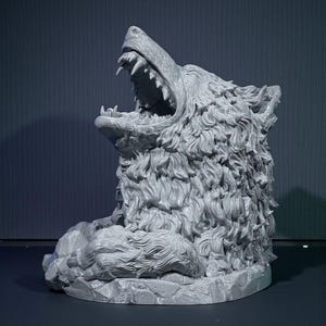 May include: A gray, detailed sculpture of a snarling wolf's head. The wolf's mouth is wide open, revealing sharp teeth. The sculpture has a textured, fur-like appearance and sits on a rocky base. The background is a dark gray.
