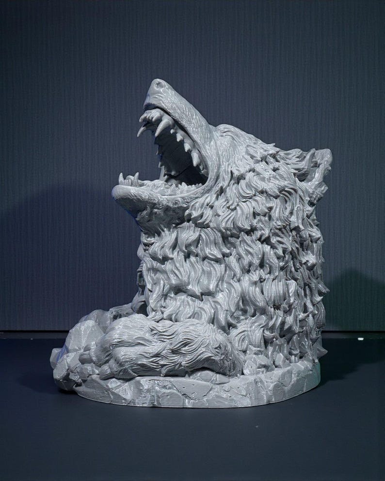 May include: A gray, detailed sculpture of a snarling wolf's head. The wolf's mouth is wide open, revealing sharp teeth. The sculpture has a textured, fur-like appearance and sits on a rocky base. The background is a dark gray.