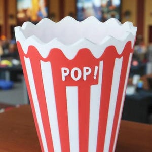 May include: A red and white striped popcorn container with the word "POP!" in white letters. The container has a scalloped white rim and is sitting on a wooden surface. The design is reminiscent of classic movie theater popcorn buckets.