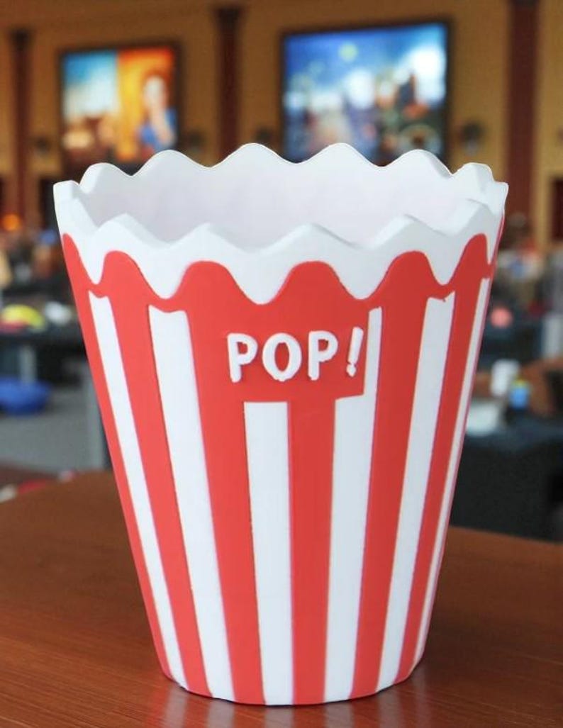 May include: A red and white striped popcorn container with the word "POP!" in white letters. The container has a scalloped white rim and is sitting on a wooden surface. The design is reminiscent of classic movie theater popcorn buckets.