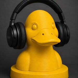 May include: A yellow duck-shaped headphone stand with black headphones. The stand is a bright yellow color and has a textured surface. The headphones are black and have the brand name "SONY" printed on the side.