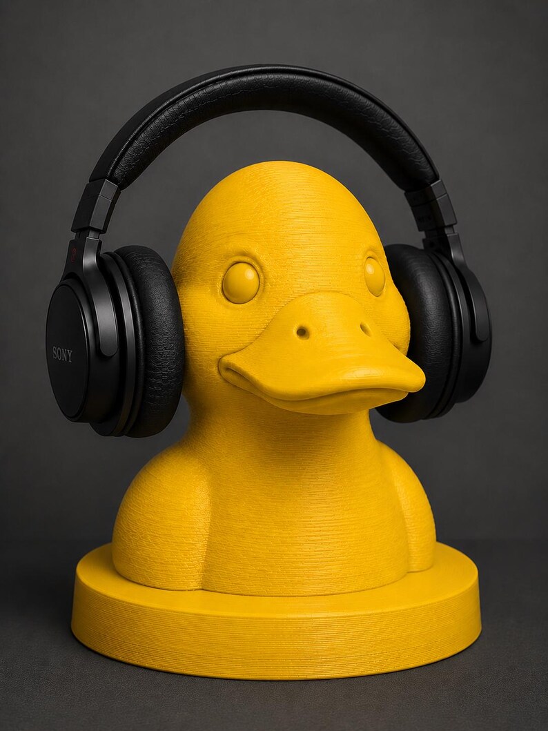 May include: A yellow duck-shaped headphone stand with black headphones. The stand is a bright yellow color and has a textured surface. The headphones are black and have the brand name "SONY" printed on the side.