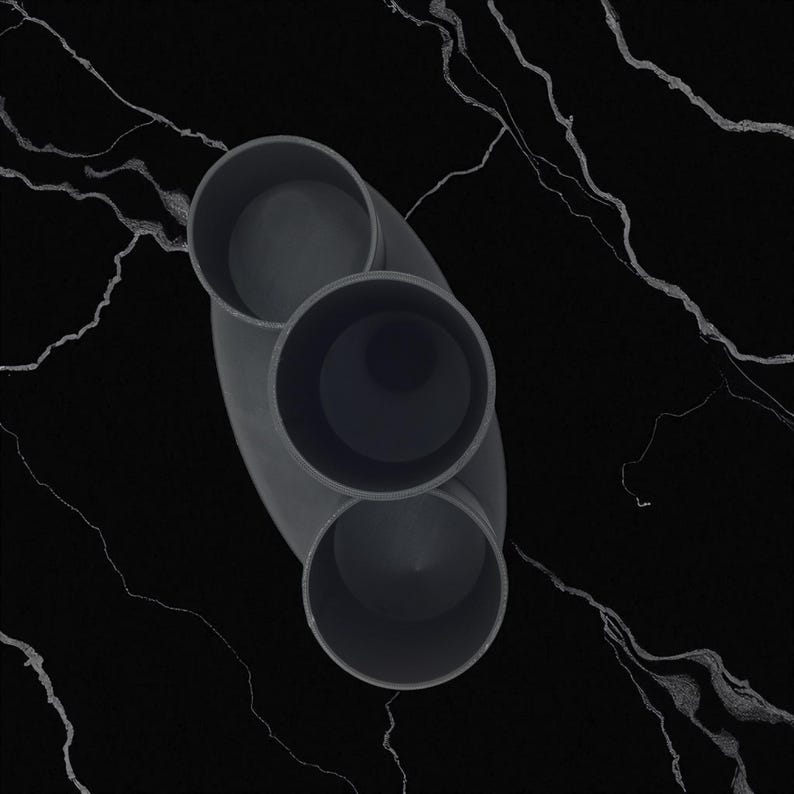 Op de afbeelding: Een donkergrijs, ovaalvormig object met drie cilindrische openingen. Het object staat tegen een zwarte achtergrond met witte adering, wat een contrast cre&euml;ert. Het ontwerp is minimalistisch en modern.