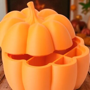 May include: An orange pumpkin-shaped container with a lid, perfect for Halloween or fall decor. The container is divided into sections, and the lid is slightly ajar, revealing the interior. The pumpkin has a stem and a textured surface.
