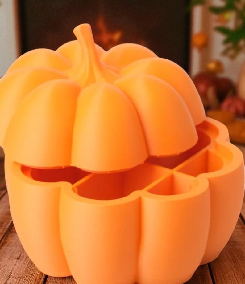 May include: An orange pumpkin-shaped container with a lid, perfect for Halloween or fall decor. The container is divided into sections, and the lid is slightly ajar, revealing the interior. The pumpkin has a stem and a textured surface.