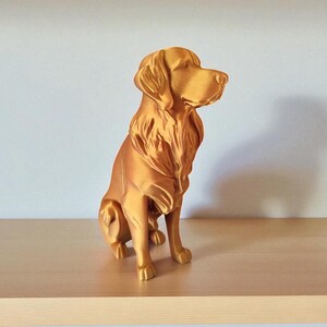 Op de afbeelding: Een goudkleurig hondenbeeld in zittende positie. De sculptuur heeft een gedetailleerde vachttextuur en kijkt naar rechts. Het hondenbeeld staat op een lichtgekleurd houten oppervlak.