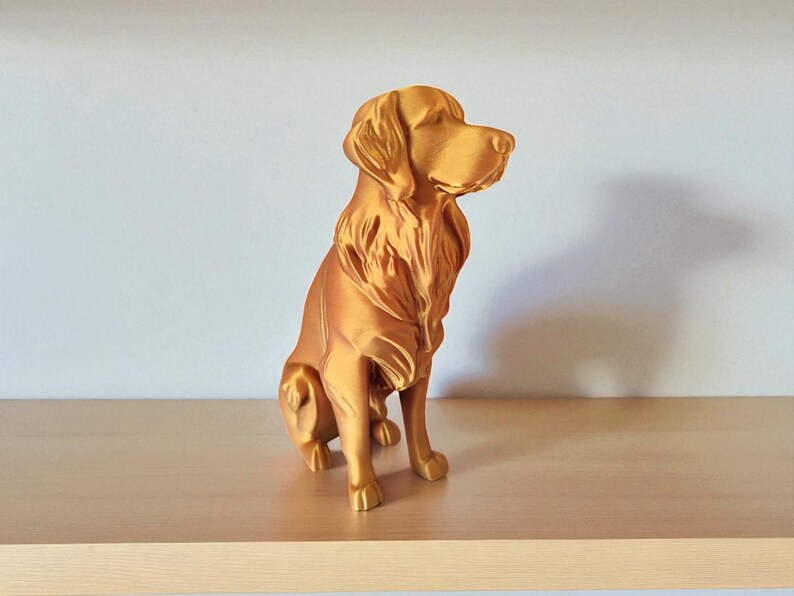 Op de afbeelding: Een goudkleurig hondenbeeld in zittende positie. De sculptuur heeft een gedetailleerde vachttextuur en kijkt naar rechts. Het hondenbeeld staat op een lichtgekleurd houten oppervlak.