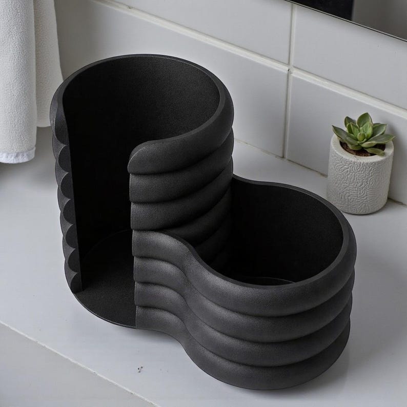 Pode incluir: Um organizador de casa de banho preto e moderno com um design ondulado &uacute;nico. O organizador tem dois compartimentos e uma superf&iacute;cie texturizada. &Eacute; feito de um material escuro e s&oacute;lido e assenta numa superf&iacute;cie branca. Uma pequena planta suculenta em vaso &eacute; vis&iacute;vel no fundo.