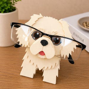 May include: A cream-colored dog-shaped eyeglass holder with black-framed glasses. The holder has a textured, layered design, with large brown eyes and a pink tongue. The glasses are positioned as if the dog is wearing them.