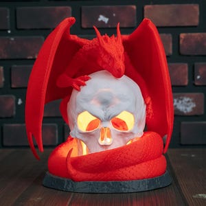 May include: A red dragon figurine perched atop a white skull with glowing yellow eyes and mouth. A red snake coils around the base. The dragon has outstretched wings. The base is dark gray.