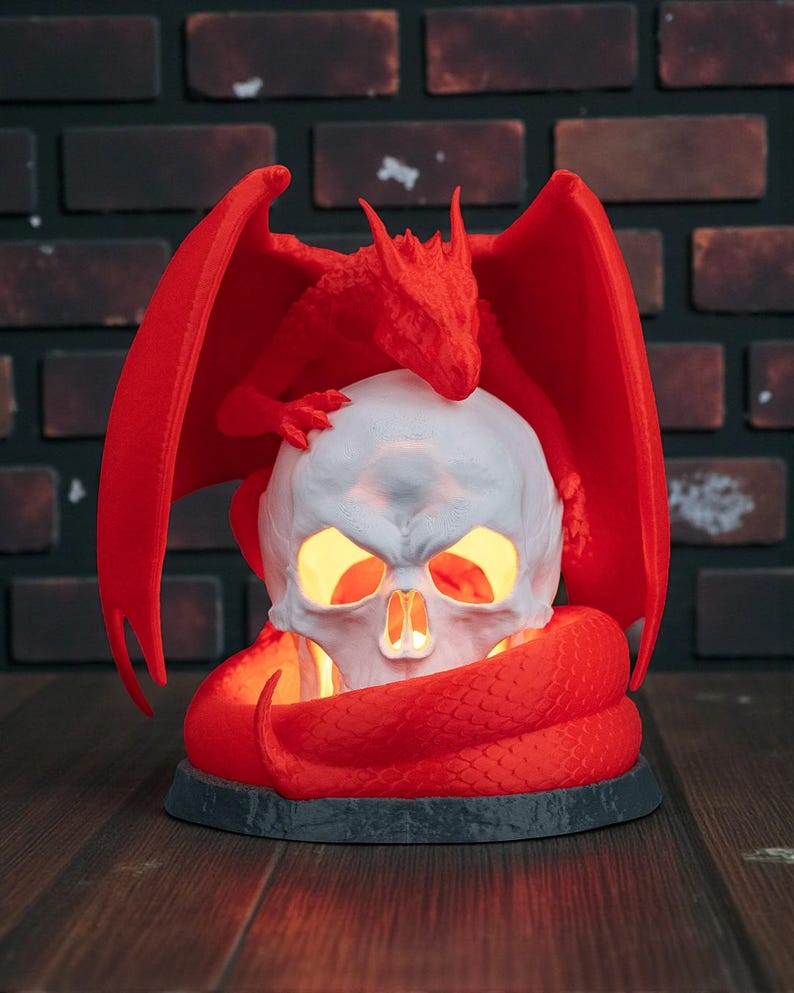 May include: A red dragon figurine perched atop a white skull with glowing yellow eyes and mouth. A red snake coils around the base. The dragon has outstretched wings. The base is dark gray.