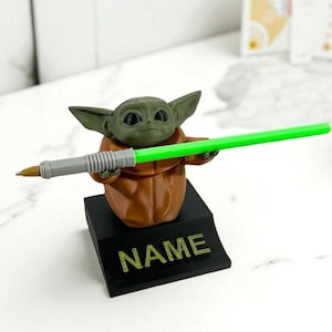 May include: A Grogu figure in green and brown, holding a green pen, mounted on a black base. The base has the word "NAME" printed on it. The pen has a grey barrel and a gold tip. The figure is a stylized representation of the character from the Star Wars franchise.