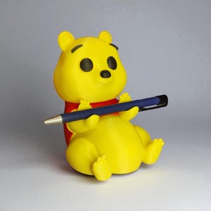 May include: A yellow Winnie the Pooh figurine holding a blue pen. The figurine has black eyes and a red shirt. The pen is held in a writing position, suggesting the figurine is writing. The background is a solid gray.