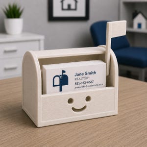 May include: A beige business card holder in the shape of a mailbox, featuring a smiling face design. It holds white business cards with "Jane Smith, REALTOR" and contact details. A small flag is attached to the side.