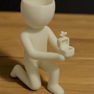 May include: A white figurine of a person kneeling, holding a small box with a butterfly on top. The figurine has a large, open head and is positioned on a wooden surface. The box is open, suggesting a ring or small item inside.